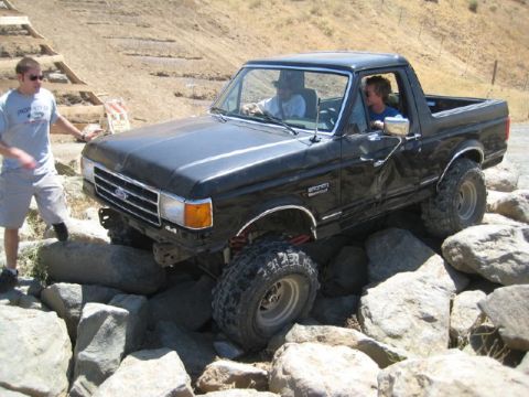 1989 Ford Bronco - A black Ford Bronco navigates a rocky obstacle course with two people inside and one person observing.