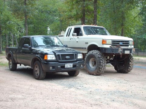1990 Ford F-150 - A black Ford Ranger and a lifted white Ford F-Series truck are parked side-by-side on a dirt lot.