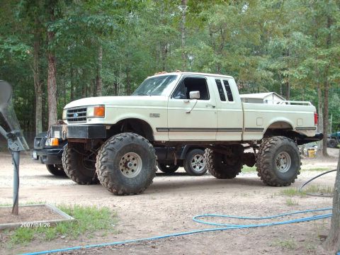 1990 Ford F-150 - A modified 1990 Ford F-150 pickup truck is shown with a lifted suspension and massive tires, parked on a dirt lot.