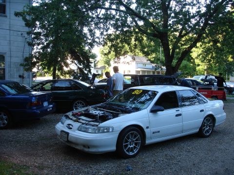 1993 Ford Taurus - Several cars are parked outside on a gravel lot, with trees overhead and houses in the background.