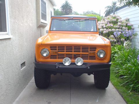 1977 Ford Bronco - This orange 1977 Ford Bronco is parked in a driveway next to a house and some flowering bushes.