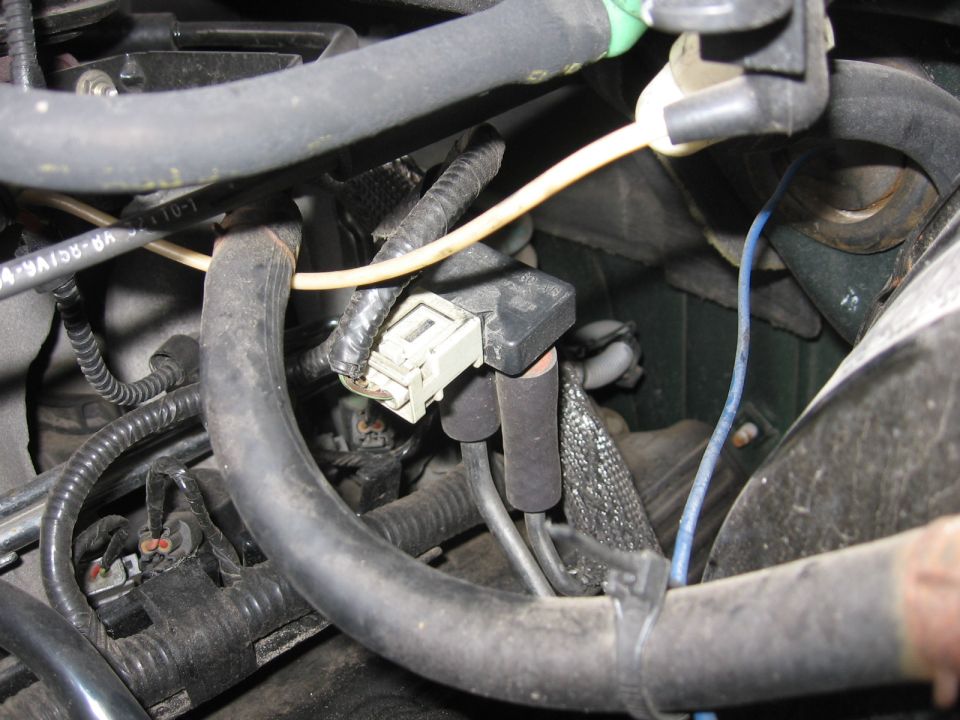 Close-up of a black sensor with a grey electrical connector attached to engine hoses and wiring