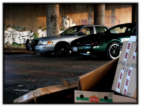 1998 Ford Crown Victoria - Two cars, a 1998 Ford Crown Victoria and a green Ford Mustang, are parked under a bridge with graffiti.