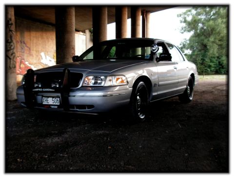 1998 Ford Crown Victoria - This silver sedan, likely a Ford Crown Victoria, is parked under a concrete overpass with graffiti on the wall.