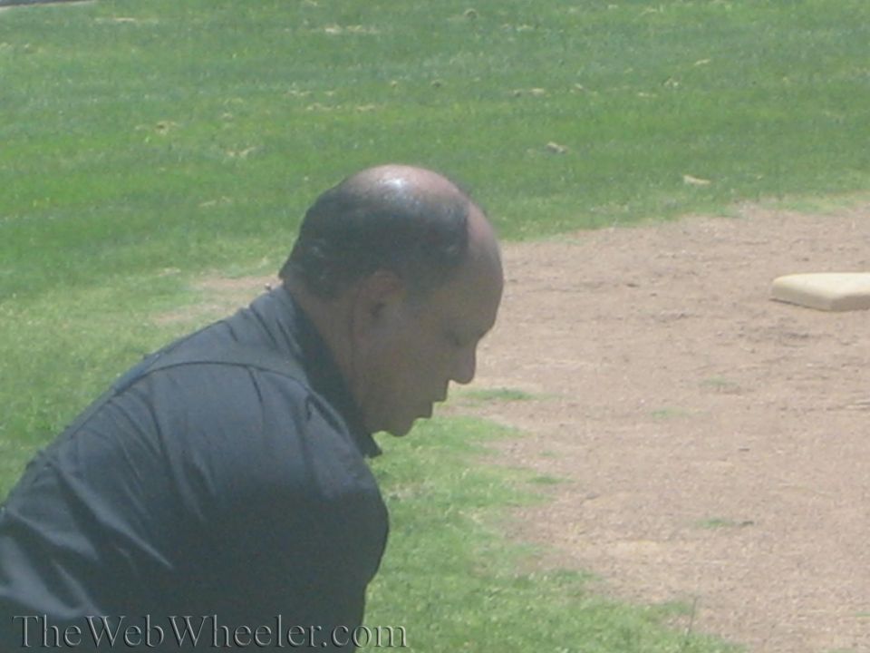 Bald man in a black shirt crouching on a grassy field, viewed in profile
