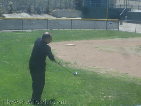 1990 Ford Bronco - man in black follows through a golf swing, club low, on a grass field with a fenced outfield behind.