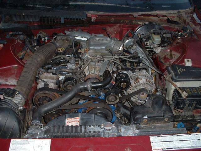 Engine bay with V6 engine, hoses, belts, and alternator visible. Some labels and text on parts