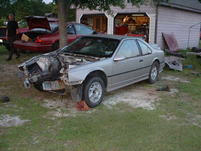 Silver Ford Thunderbird coupe with front clip and hood removed, being parted out in a yard