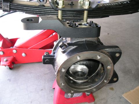 1986 Toyota Hilux - Close-up of a bare solid front axle housing end resting on a red floor jack, with a black powder-coated spring-over perch/shock-mount brack…