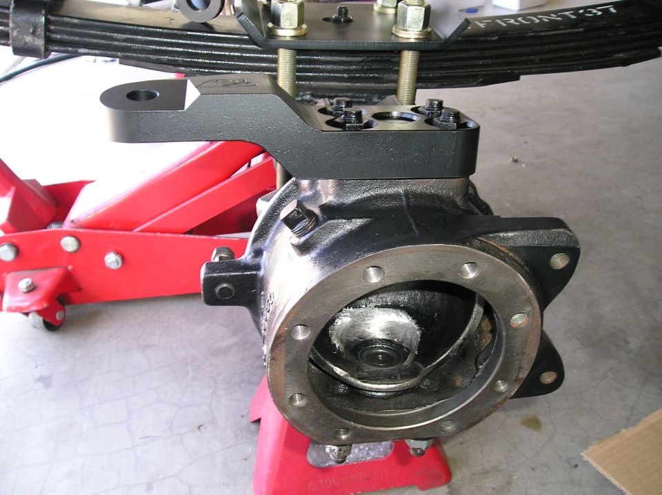 Front axle housing end with black spring-over perch bracket clamped to leaf pack on a red floor jack