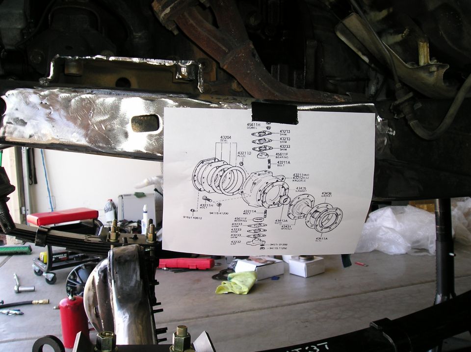 Toyota knuckle/spindle parts diagram taped to a stripped frame rail during a Hilux front axle rebuild in a garage