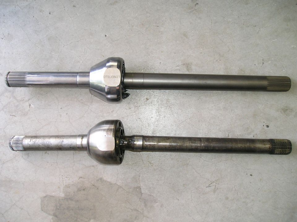 New Longfield front axle shaft with CV joint next to a used stock axle shaft on concrete floor