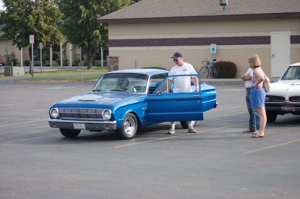 Blue classic car with driver's door open, man standing by it. Another car is parked to the right