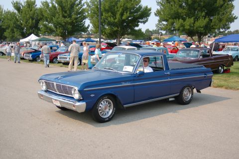 1967 Ford Ranchero - A blue pickup truck is parked at an outdoor event with other cars and people milling around.