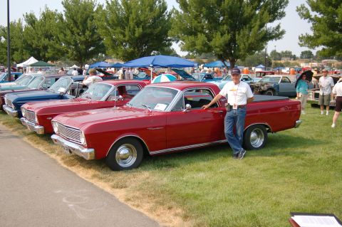 1967 Ford Ranchero - A man stands next to a red 1967 Ford Ranchero at a car show, with other vehicles lined up in the background.