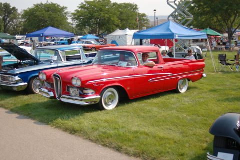 1967 Ford Ranchero - A red pickup truck with distinctive chrome accents and whitewall tires is parked on a grassy field at an outdoor event.