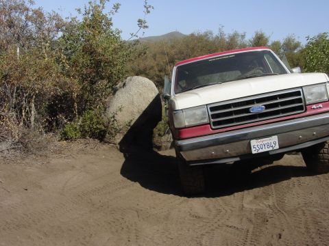 1994 Ford Bronco - This two-tone Ford Bronco is shown off-roading on a dirt path with a large boulder to its left.