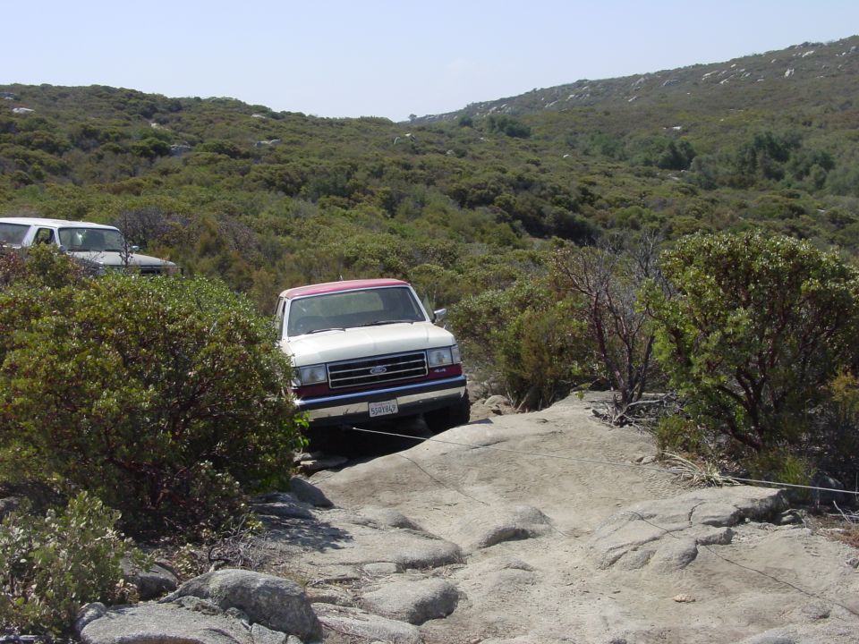 White Ford Bronco with red roof driving over rocks on a trail. Another SUV is visible in the background