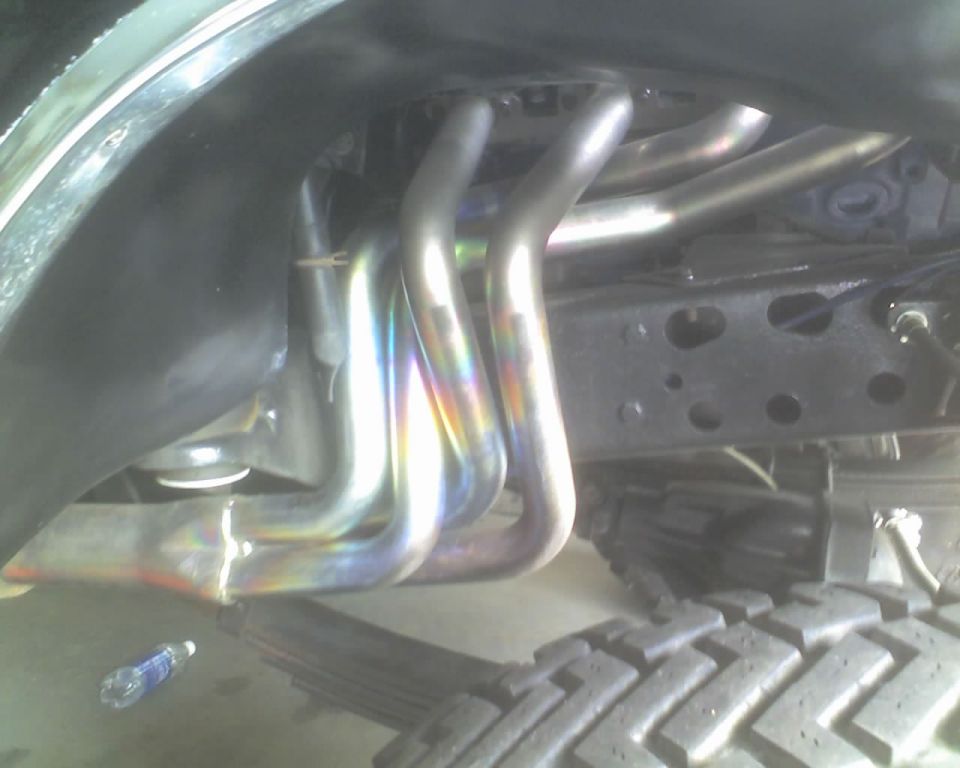 Exhaust headers on a vehicle frame, with a tire and leaf spring visible. A water bottle is on the ground