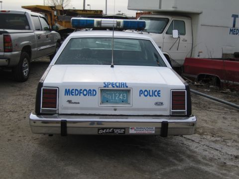 1991 Ford LTD Crown Victoria - A police car with a light bar sits in a junkyard next to other vehicles and debris.
