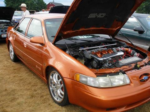 1997 Ford Taurus - A custom metallic orange Ford Taurus SHO sedan shown at an outdoor car show with its hood open, revealing a modified engine bay including a…