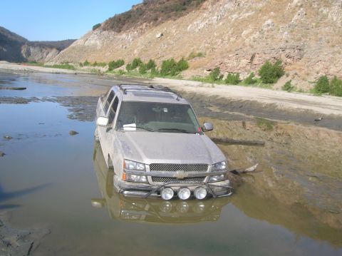 1985 Ford Bronco - A silver SUV is partially submerged in muddy water, with its front end facing the camera and off-road lights visible.