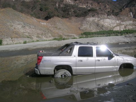 1985 Ford Bronco - Silver pickup truck submerged in muddy water, stuck in a riverbed.