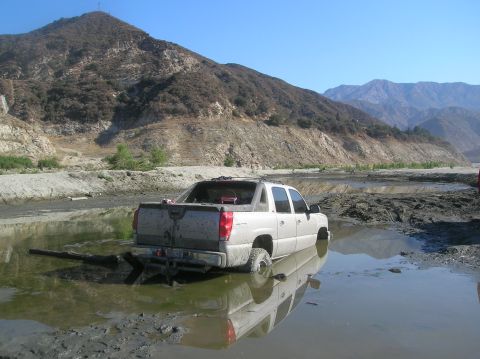 1985 Ford Bronco - A pickup truck is submerged in muddy water, with its tires sunk deep into the mire.