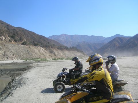 1985 Ford Bronco - Riders on ATVs traverse a dry riverbed, with mountainous terrain under a clear blue sky.