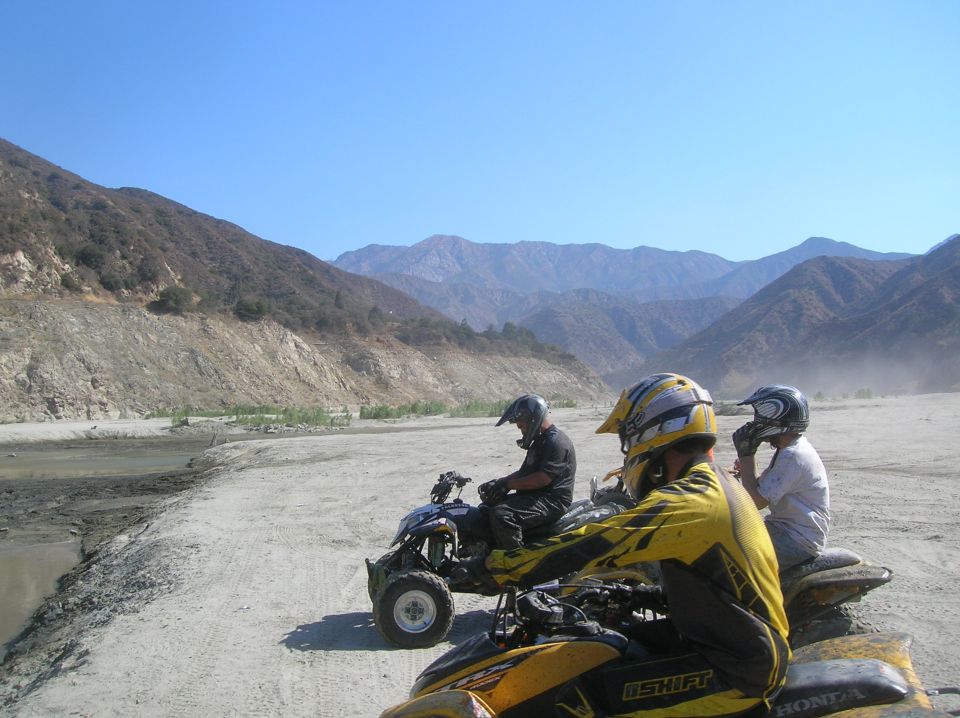 Three ATVs with riders in a dry riverbed with mountains in the background