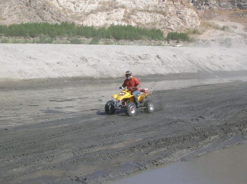 1985 Ford Bronco - A person rides a yellow ATV on a muddy track with a sandy embankment in the background.