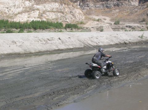 1985 Ford Bronco - ATV rider navigates a muddy terrain in a dry riverbed with arid hills.