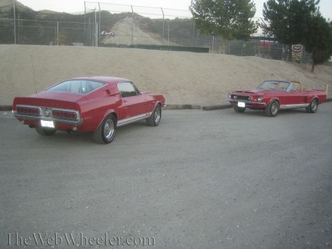 1990 Ford Bronco - red Mustang fastback and a red Mustang convertible sit parked side by side on gravel beside a dirt embankment.