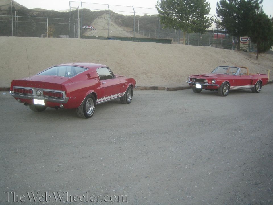 Red Mustang fastback and red Mustang convertible parked on gravel by a dirt hill