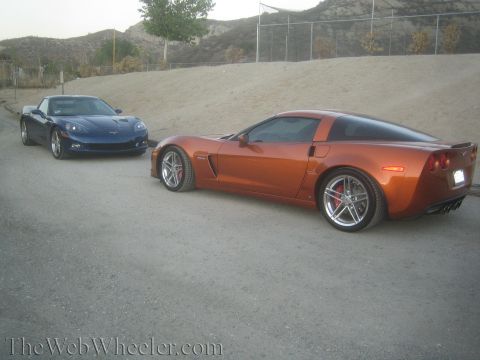 1990 Ford Bronco - orange Corvette coupe and a blue Corvette sit parked on gravel in front of a bare dirt embankment.