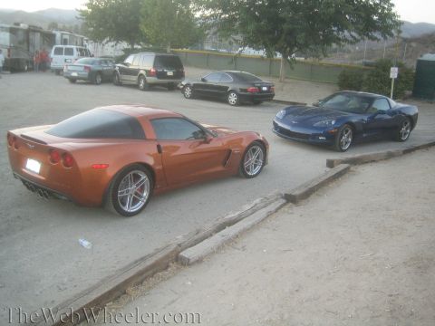 1990 Ford Bronco - orange Corvette and a blue Corvette sit parked in a gravel lot among sedans and a van near hillsides at dusk.