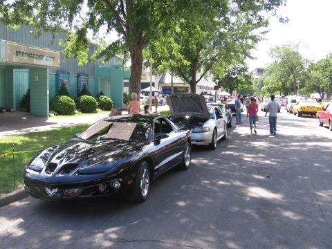 1989 Ford Mustang - Two cars are parked on a street during a car show event with people around.