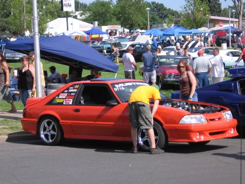 1989 Ford Mustang - A person inspects an orange coupe at a car show, with other vehicles and tents in the background.
