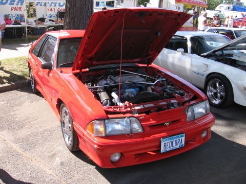 1989 Ford Mustang - A red 1989 Ford Mustang is displayed with its hood open, revealing the engine bay, parked next to a white Pontiac Trans Am.