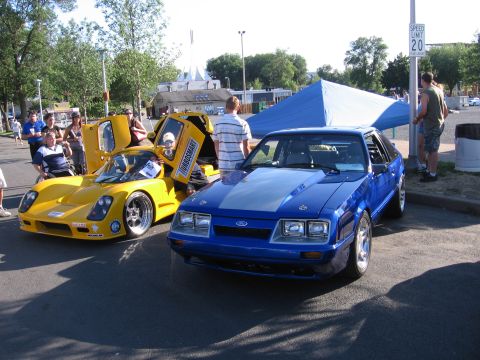 1989 Ford Mustang - Two cars, a blue Ford Mustang and a yellow sports car, parked outside on a sunny day.