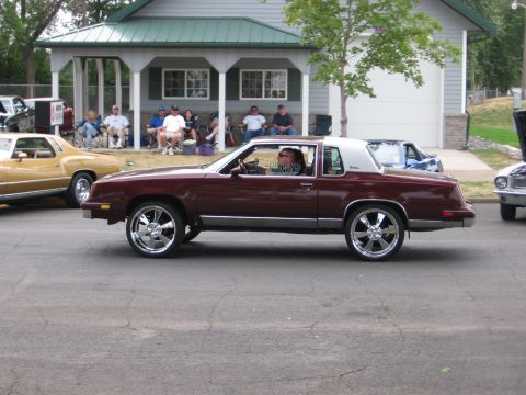 1989 Ford Mustang - A maroon coupe with a white roof drives down the street, while spectators watch from a porch.