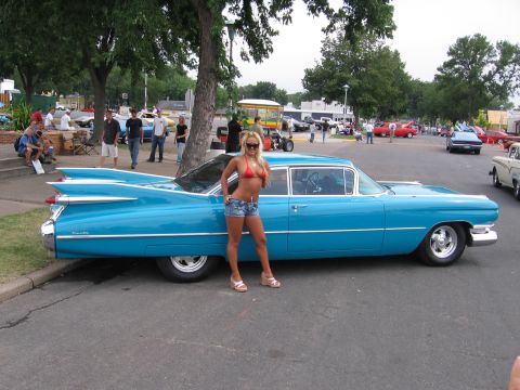 1989 Ford Mustang - A woman poses next to a bright blue 1959 Cadillac sedan with distinctive tail fins at an outdoor car show.