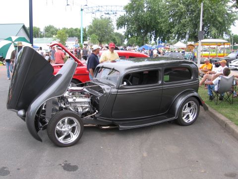 1989 Ford Mustang - A dark gray hot rod sedan is displayed with its hood open, revealing a chrome engine and wheels.