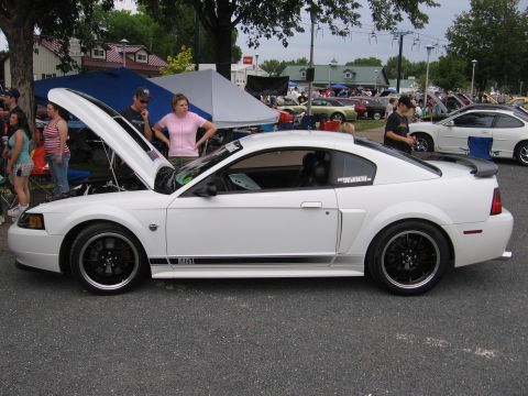1989 Ford Mustang - A white Ford Mustang is displayed with its hood up at an outdoor event, with spectators gathered around.