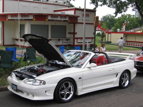 1989 Ford Mustang - A white convertible sports car, likely a 1994 Ford Mustang, is parked with its hood open in front of a building with red trim.