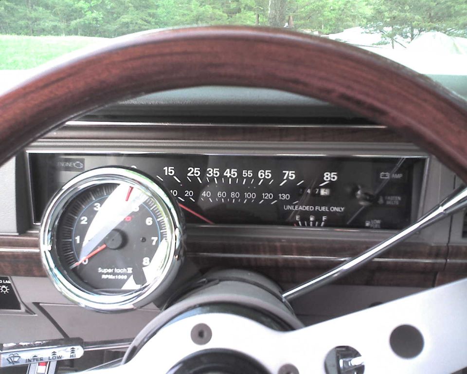 Car dashboard cluster with speedometer and add-on chrome Super Tach II tachometer, seen through woodgrain steering wheel.