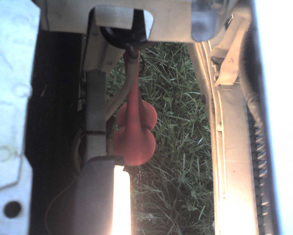 Red rubber bulb component and metal fitting on tan panel over grass, viewed through vehicle opening