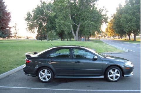 2005 Mazda 6 - A dark gray 2005 Mazda 6 sedan is parked in a lot with trees and a road in the background.