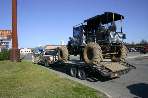 2003 Jeep TJ - A custom monster truck with oversized tires is loaded onto a flatbed trailer, being towed by a pickup truck.