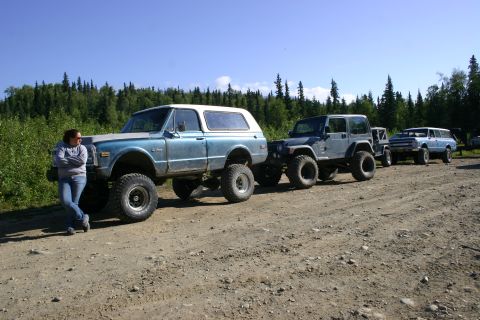 2003 Jeep TJ - Three lifted off-road vehicles parked on a dirt road with a wooded background.